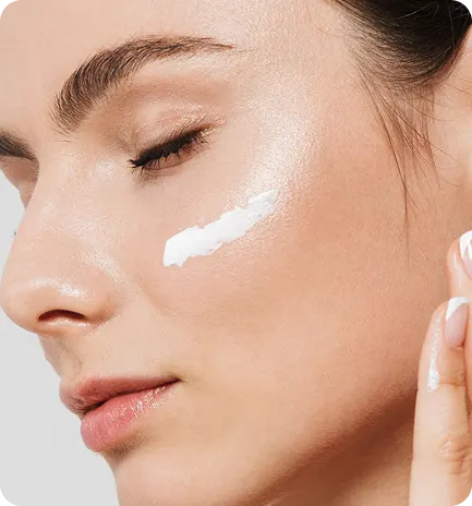 Close-up of a woman applying cream to her face with a neutral background